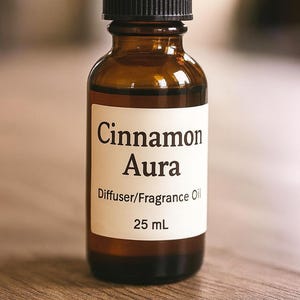 May include: A brown glass bottle with a black cap, labeled "Cinnamon Aura Diffuser/Fragrance Oil 25 mL." The bottle sits on a wooden surface. The label has a white background with black text.