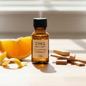 May include: A small amber glass bottle with a black cap, labeled "ZING AND ZEST ALL NATURAL FRAGRANCE 3 mL", sits next to an orange slice and wood pieces. Orange peel curls are in the foreground. The scene is lit by natural light.
