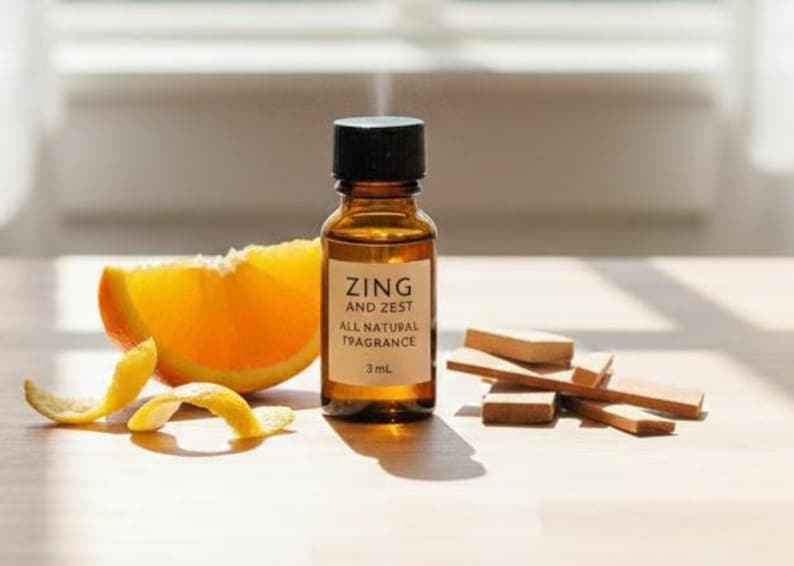 May include: A small amber glass bottle with a black cap, labeled "ZING AND ZEST ALL NATURAL FRAGRANCE 3 mL", sits next to an orange slice and wood pieces. Orange peel curls are in the foreground. The scene is lit by natural light.