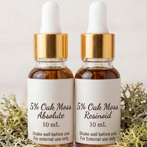 May include: Two glass dropper bottles with gold collars and white tops. Each bottle has a white label with black text that reads "5% Oak Moss Absolute 10 mL" and "5% Oak Moss Resinoid 10 mL". The text "Shake well before use. For external use only" is also printed on the labels.