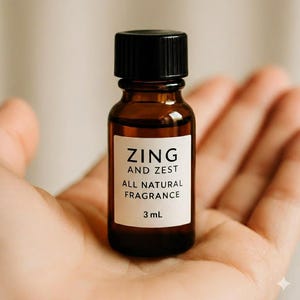 May include: A small amber glass bottle with a black cap, labeled "ZING AND ZEST ALL NATURAL FRAGRANCE" and 3 mL. The bottle is held in an open hand, suggesting a focus on natural fragrance products.