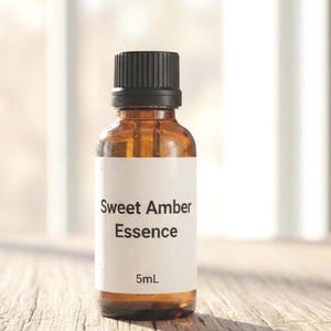 May include: A small amber glass bottle with a black cap. The bottle has a white label with the words "Sweet Amber Essence" and "5mL" printed on it. The bottle is sitting on a wooden surface.