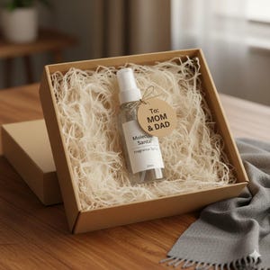 Puede incluir: Un frasco de spray de fragancia transparente con tapa y etiqueta blancas, presentado en una caja de regalo marr&oacute;n llena de papel triturado. La etiqueta dice "Molecular Santal Fragrance Spray 25ml". Una etiqueta redonda atada a la botella dice "To: MOM & DAD."