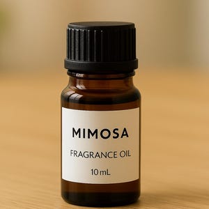 May include: A small, amber-colored glass bottle of Mimosa fragrance oil with a black cap. The white label displays the product name and volume, 10 mL. The bottle is on a light brown wooden surface.