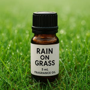 May include: A small brown glass bottle with a black cap, labeled "RAIN ON GRASS 5 mL FRAGRANCE OIL." The bottle is set against a backdrop of vibrant green grass with water droplets, creating a fresh, natural aesthetic.
