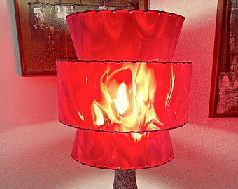 May include: A three-tiered red lampshade with a textured, gray ceramic base. The lampshade has a rippled, translucent effect, and the base has a swirling pattern.