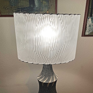 May include: A mid-century modern table lamp with a white pleated lampshade and a black and gold ceramic base with a swirling pattern.