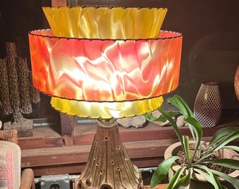 May include: A vintage table lamp with a brown ceramic base featuring a textured design. The lamp has two shades, the bottom one is red and yellow, and the top one is yellow. The lamp is lit, and the shades are translucent.