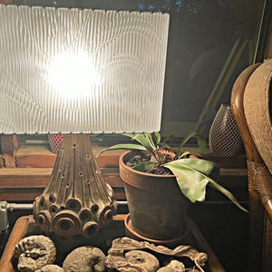 May include: A vintage lamp with a white textured shade and a brown ceramic base with a unique design. The lamp is on a table with a potted plant and some decorative rocks.