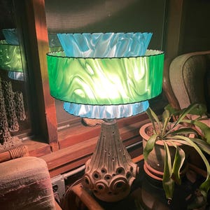 May include: A vintage table lamp with a layered lampshade in shades of blue and green. The lampshade has a wavy, marbled design. The base of the lamp is a textured, earthy brown color with circular cutouts. The lamp is placed near a window.