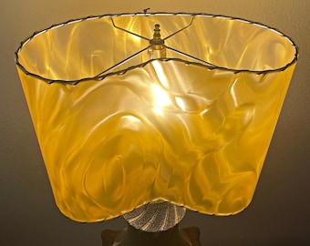 May include: A vintage table lamp with a large, yellow, textured lampshade. The shade has a wavy design and is supported by a brass frame. The base is a cream color with a textured pattern. The lamp is turned on, emitting a warm glow.