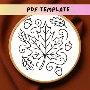Puede incluir: Un bastidor de bordado blanco con un dibujo lineal negro de una hoja de arce, bellotas y otras hojas de otoño. Las palabras "PDF TEMPLATE" están en la parte superior. El bastidor está sobre un fondo marrón.