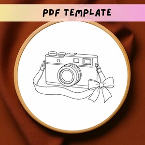 May include: A circular wooden embroidery hoop containing a white fabric panel with a black line art illustration of a camera featuring a decorative bow on the strap. The text PDF TEMPLATE is printed in black at the top of the image.