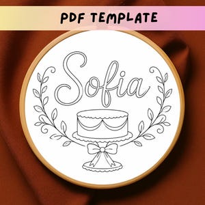 May include: A circular wooden embroidery hoop containing a white fabric panel with a black line art pattern. The design features the name Sofia in cursive script above a cake on a pedestal, framed by two symmetrical leafy vine branches.