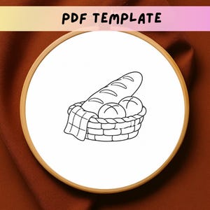 Può includere: Un telaio da ricamo circolare in legno contenente tessuto bianco con un'illustrazione a linee nere di un cestino pieno di una baguette e due pagnotte rotonde. Il testo PDF TEMPLATE è stampato in nero nella parte superiore dell'immagine.