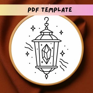 May include: A black and white line drawing of a lantern with a crystal inside, surrounded by stars and wavy lines, set in a wooden embroidery hoop. The text "PDF TEMPLATE" is at the top.