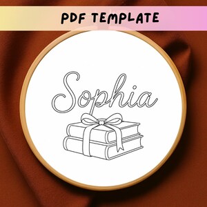 May include: A circular wooden embroidery hoop containing white fabric with a black line art design. The design features the name Sophia in cursive script above a stack of two books tied together with a ribbon bow. The text PDF TEMPLATE is visible.