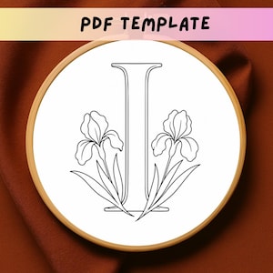 May include: A PDF template featuring a black and white line art illustration of the capital letter I centered within a circular wooden embroidery hoop. The letter is flanked by two iris flowers with leaves on either side, set against a white background.