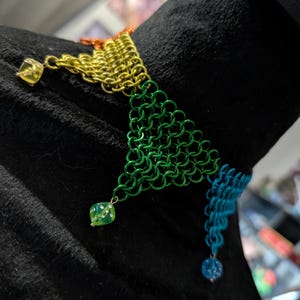 May include: A colorful chainmail necklace with a triangular design. The necklace features gold, green, and blue sections, with small dice and bead charms. The chainmail is made of small, interlocking metal rings.