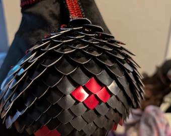 May include: Close-up of a black and red chainmail shoulder pad. The shoulder pad is made of small, interlocking metal scales. The scales are arranged in a pattern that resembles a dragon's scales. The shoulder pad is accented with red leather straps.