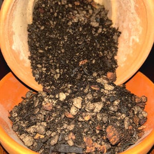 May include: An orange ceramic pot is tilted over a matching bowl, spilling dark potting soil. The soil is a mix of dark brown, tan, and light brown materials, including small rocks and wood chips. The scene is lit with warm, natural light.