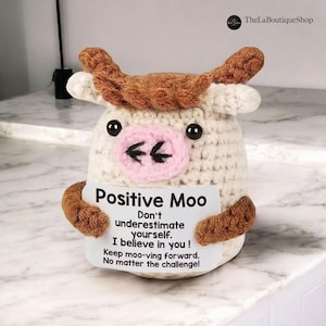 Puede incluir: Figura de vaca de ganchillo con cuerpo blanco, cuernos y brazos marrones y nariz rosa. La vaca sostiene un letrero que dice "Positive Moo" y un mensaje inspirador. La figura está sobre una superficie de mármol.