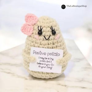 Puede incluir: Una figura de patata de ganchillo en color beige claro, con un lazo rosa y ojos negros. La patata sostiene una tarjeta que dice "Positive potato" y un mensaje motivador. Hecho a mano.