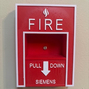 May include: A red fire alarm pull station with white text reading "FIRE" and "PULL DOWN". The word "SIEMENS" is also printed on the alarm. The alarm is mounted on a beige wall.