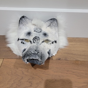 May include: A white and gray wolf mask with black markings around the eyes and nose. The mask has a fluffy white fur mane and decorative markings above the eyes. The mask is lying on a wooden surface.