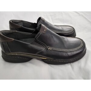 May include: A pair of black leather slip-on shoes with white stitching and brown accents. The shoes have a rounded toe and a thick sole. The stitching is visible around the edges and on the top of the shoe.