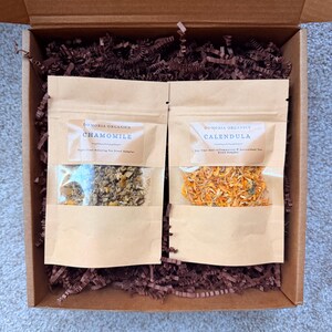 Puede incluir: Una caja de regalo con dos bolsas de papel kraft de t&eacute; de hierbas. Una bolsa est&aacute; etiquetada como "Chamomile" y la otra como "Calendula". La caja est&aacute; llena de papel arrugado marr&oacute;n. La bolsa de manzanilla contiene flores secas. La bolsa de cal&eacute;ndula contiene p&eacute;talos naranjas.