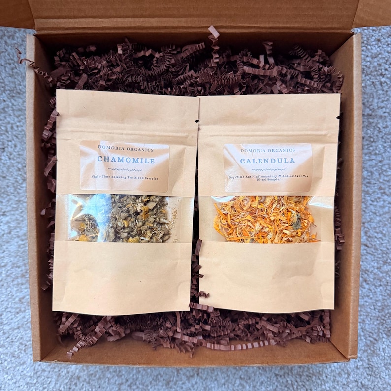 Puede incluir: Una caja de regalo con dos bolsas de papel kraft de t&eacute; de hierbas. Una bolsa est&aacute; etiquetada como "Chamomile" y la otra como "Calendula". La caja est&aacute; llena de papel arrugado marr&oacute;n. La bolsa de manzanilla contiene flores secas. La bolsa de cal&eacute;ndula contiene p&eacute;talos naranjas.