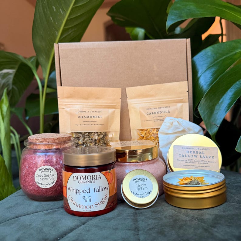 Puede incluir: Una colecci&oacute;n de productos Domoria Organics, que incluye frascos de sal del Mar Muerto, sebo batido y b&aacute;lsamo labial, junto con bolsas de manzanilla y cal&eacute;ndula. Los art&iacute;culos est&aacute;n dispuestos sobre una superficie verde.