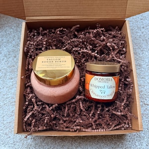 Puede incluir: Una caja de regalo con dos tarros de Domoria Organics. Uno es un exfoliante de az&uacute;car de sebo rosa con tapa dorada. El otro es Whipped Tallow Cinnamon Sugar con una etiqueta naranja y tapa dorada. La caja est&aacute; llena de papel marr&oacute;n.