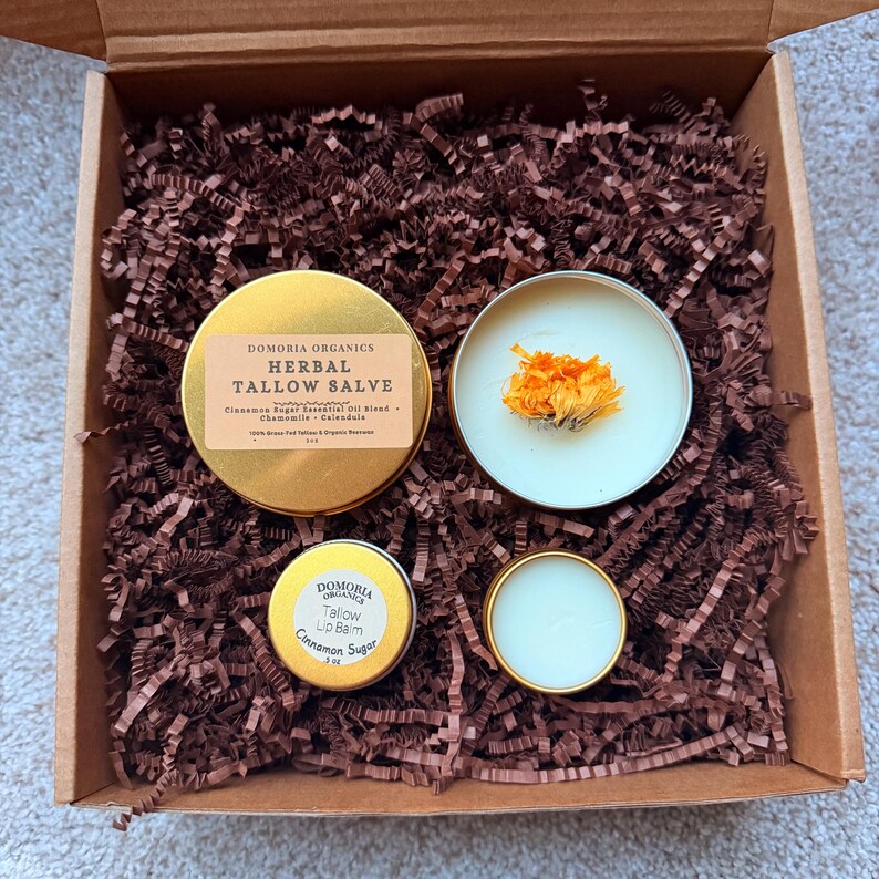 Puede incluir: Una caja de regalo llena de virutas de papel marr&oacute;n, que contiene cuatro latas doradas. Dos latas est&aacute;n abiertas, revelando un b&aacute;lsamo blanco y flores naranjas secas. Las otras dos latas tienen etiquetas que dicen "Domoria Organics Herbal Tallow Salve" y "Tallow Lip Balm Cinnamon Sugar".