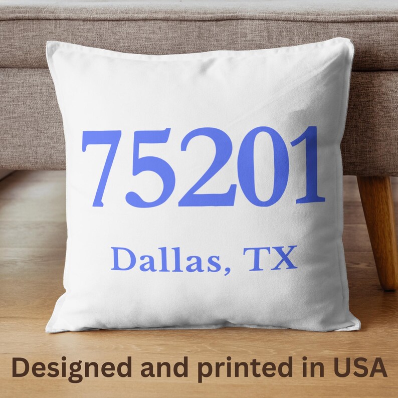 May include: White square cushion featuring the blue text "75201" above "Dallas, TX". The text "Designed and printed in USA" is at the bottom. The cushion is on a wooden surface.