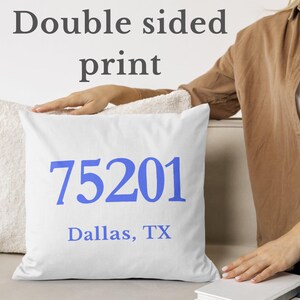 May include: White square cushion with the blue text "75201 Dallas, TX". The words "Double sided print" are above the cushion. The cushion is on a white surface.