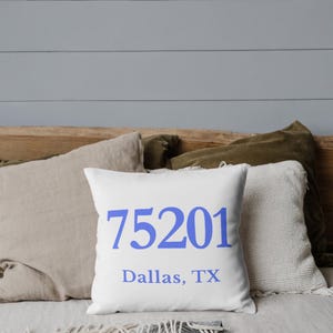 May include: White square cushion with the blue text "75201 Dallas, TX". The cushion is on a bed with other cushions in beige and green. The background is a light grey wall.