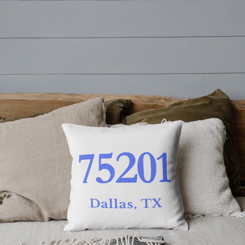 May include: White square cushion with the blue text "75201 Dallas, TX". The cushion is on a bed with other cushions in beige and green. The background is a light grey wall.