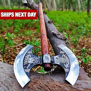 Könnte beinhalten: Eine Doppelaxt mit poliertem Stahlkopf und Holzgriff. Der Axtkopf weist aufwendige eingravierte Designs auf. Der Griff ist dunkelbraun. Die Axt liegt auf einem Ast.