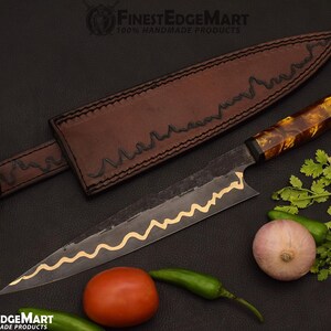 May include: A handmade chef's knife with a brown leather sheath. The knife has a dark blade with a gold wavy design and a patterned handle. Fresh vegetables, including a tomato, onion, and peppers, are arranged around the knife.