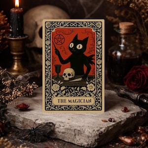 Könnte beinhalten: Eine Tarotkarte mit einem schwarzen Katzenmagier, der einen Zauberstab hält, mit einem Schädel und einem Kelch auf einem Tisch. Die Karte hat einen roten Hintergrund mit Blumen- und Sterndesigns. Der Text "THE MAGICIAN" steht unten.
