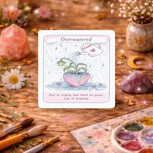 Könnte beinhalten: Eine quadratische Karte mit dem Wort "Overwatered" über einer Illustration einer Pflanze in einem rosa herzförmigen Topf, die bewässert wird. Der Text "You're trying too hard to grow. Let it breathe" steht darunter. Die Karte liegt auf einer Holzoberfläche.