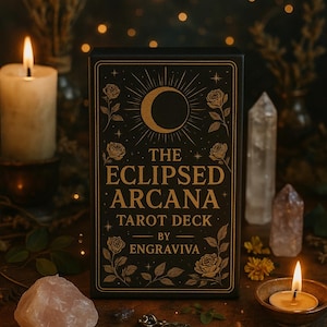 Könnte beinhalten: Eine schwarze Schachtel mit goldener Beschriftung, die "The Eclipsed Arcana Tarot Deck by Engraviva" lautet. Die Schachtel zeigt einen Halbmond und Rosen in einem goldenen Design auf schwarzem Hintergrund. Die Schachtel ist von Kerzen, Kristallen und anderen Dekorationsgegenständen umgeben.