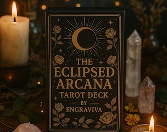 May include: A black box with gold lettering that reads "The Eclipsed Arcana Tarot Deck by Engraviva". The box features a crescent moon and roses in a gold design against a black background. The box is surrounded by candles, crystals, and other decorative items.