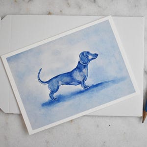Puede incluir: Una pintura de acuarela azul de un perro salchicha sobre una tarjeta blanca. La tarjeta est&aacute; colocada sobre un sobre blanco. Un l&aacute;piz azul est&aacute; al lado de la tarjeta. El fondo es un lavado azul claro.
