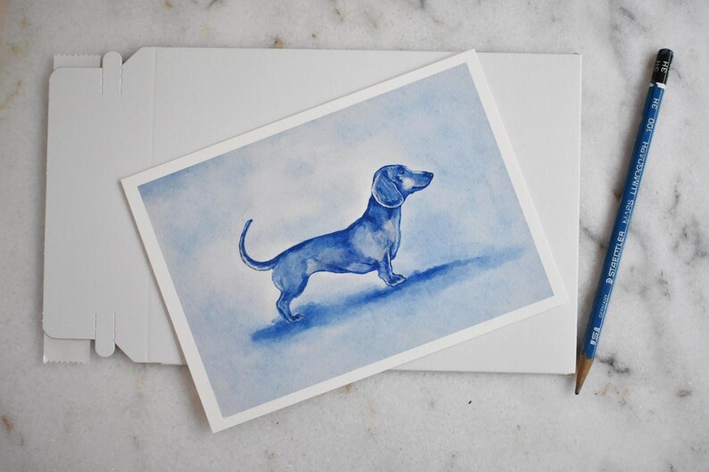 Puede incluir: Una pintura de acuarela azul de un perro salchicha sobre una tarjeta blanca. La tarjeta est&aacute; colocada sobre un sobre blanco. Un l&aacute;piz azul est&aacute; al lado de la tarjeta. El fondo es un lavado azul claro.