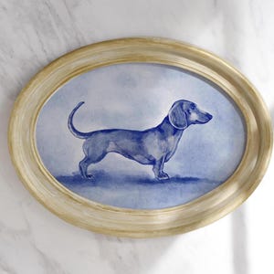 Puede incluir: Pintura de acuarela enmarcada ovalada de un perro salchicha en tonos azules. El perro est&aacute; representado de perfil, con un cuerpo largo y patas cortas. El marco es de color beige claro y el fondo es azul suave.