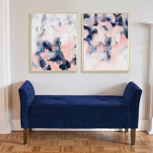 May include: Two abstract paintings in gold frames with pink, blue, and gray watercolor-like designs. The paintings are hanging on a white wall above a dark blue velvet bench.