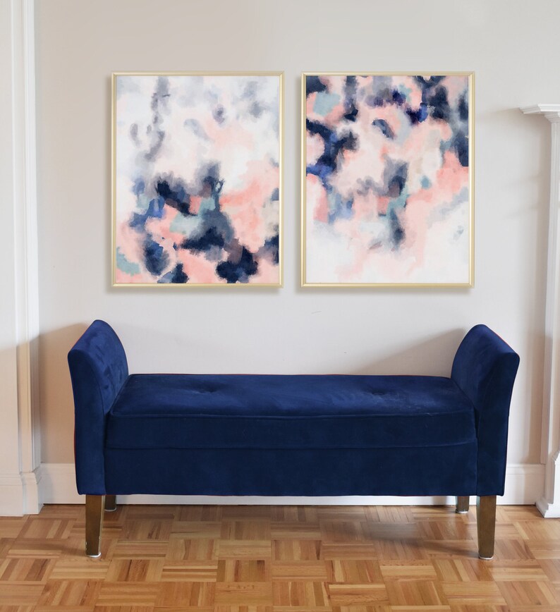 May include: Two abstract paintings in gold frames with pink, blue, and gray watercolor-like designs. The paintings are hanging on a white wall above a dark blue velvet bench.